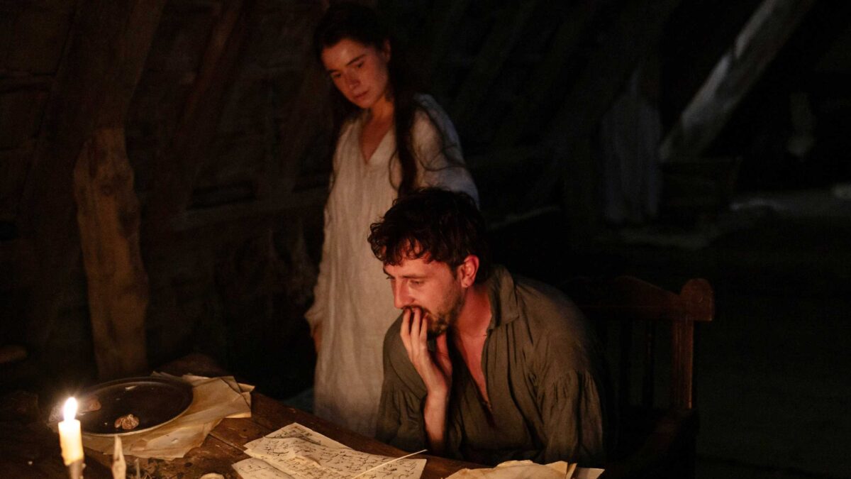 Agnes standing over Shakespeare as he sits at his candlelit desk in Hamnet