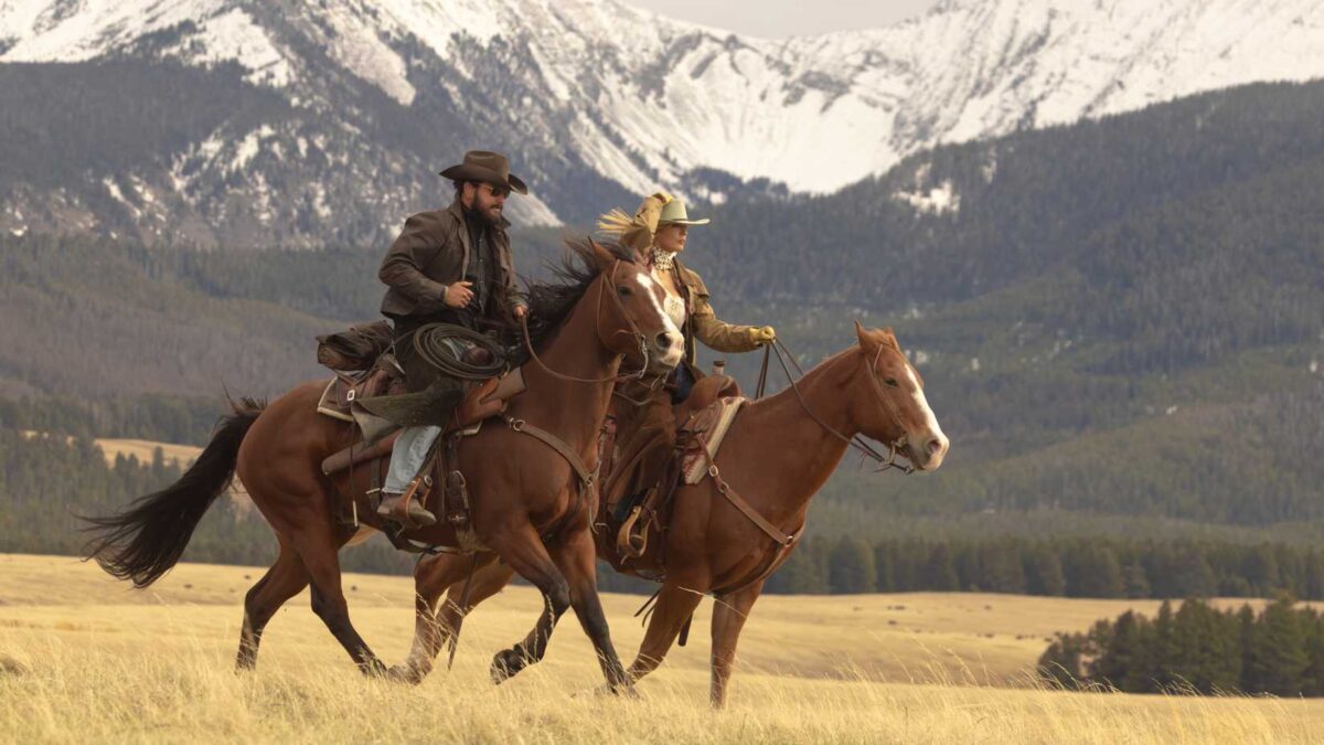 Beth and Rip ride horses through a field with mountains in the background in Yellowstone sequel series Dutton Ranch