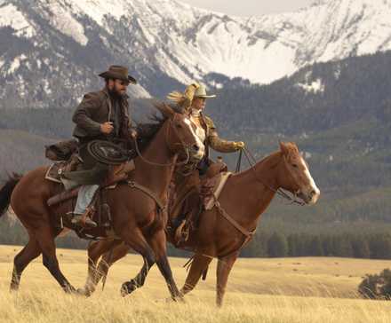 Beth and Rip ride horses through a field with mountains in the background in Yellowstone sequel series Dutton Ranch